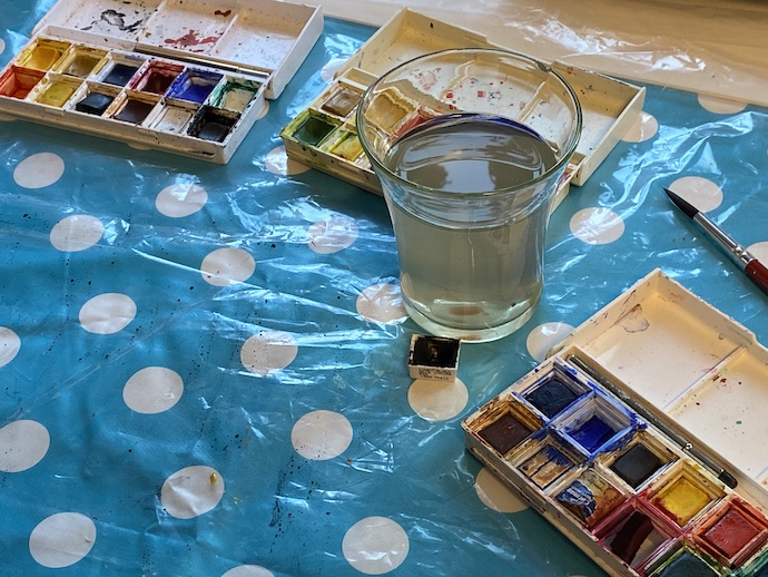 På en vitprickig vaxduk ser man paletter med akvarellfärger och ett glas med grumligt vatten